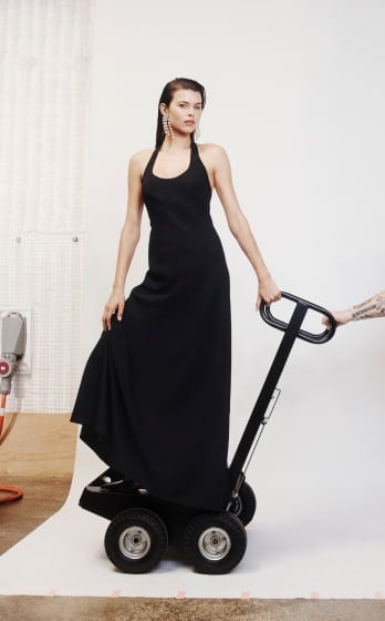 Adult woman in a black dress stands on a black motorized cart in a photo studio with a white backdrop and grey floors.