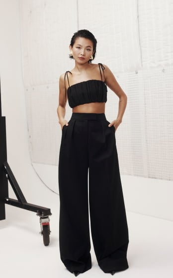 Woman in black crop top and wide-leg pants posing in photo studio. She has short dark hair and is wearing gold earrings.