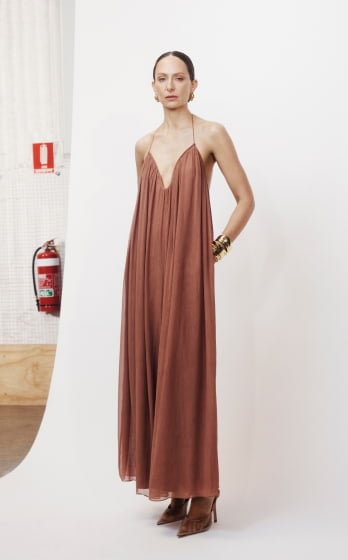 A woman models a long, flowing brown dress with a plunging neckline in a studio setting, accented by gold jewelry and brown high heels.