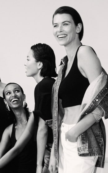 Group of smiling women in casual fashion wearing denim and tops against a white background, posing together in a studio setting.