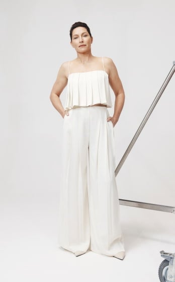 A mature woman stands in a photo studio, wearing a cream-colored, pleated two-piece outfit, posing with her hands in her pockets.