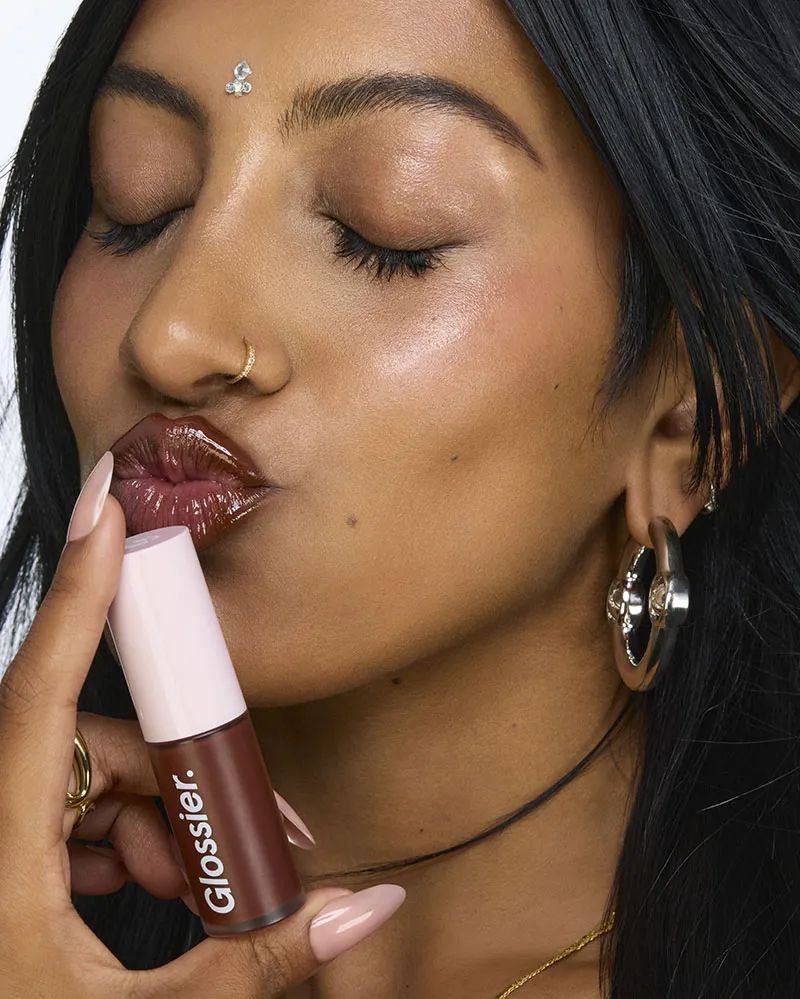 Close-up of a tan-skinned woman with a bindi, closing her eyes and holding a tube of Glossier lip gloss near her glossy lips.