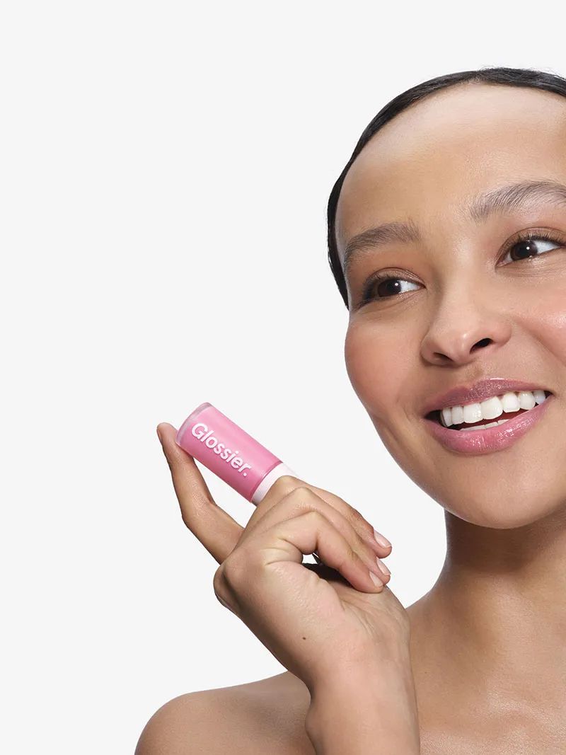 Close-up of a young woman holding a tube of pink lip gloss. The Glossier brand lip product is in focus against a plain background.
