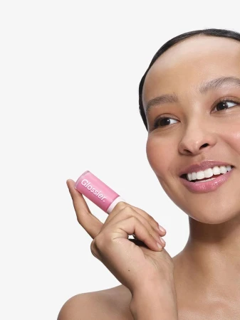 Close-up portrait of a smiling woman holding a pink Glossier lip glaze tube against a white seamless background
