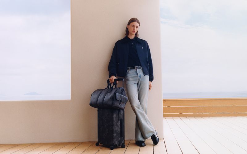 A model stands near a wall with a blue denim carry-on bag and a rolling suitcase, with a blue sky and ocean in the background.