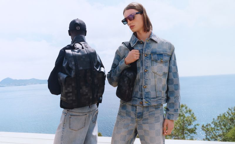 Two stylish men pose outdoors in matching patterned denim jackets and jeans. One wears a backpack and baseball cap, the other sunglasses.