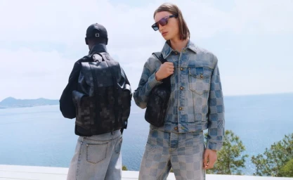 Two models outdoors by the sea wearing designer denim and carrying dark leather backpacks for Louis Vuitton.