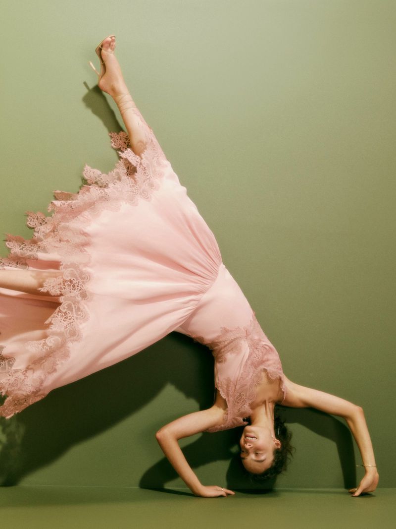 Young woman in a pink, lace-trimmed nightgown does a handstand against an olive green wall, lit by soft studio lighting.