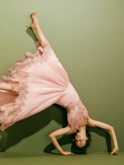 Young woman in a pink, lace-trimmed nightgown does a handstand against an olive green wall, lit by soft studio lighting.