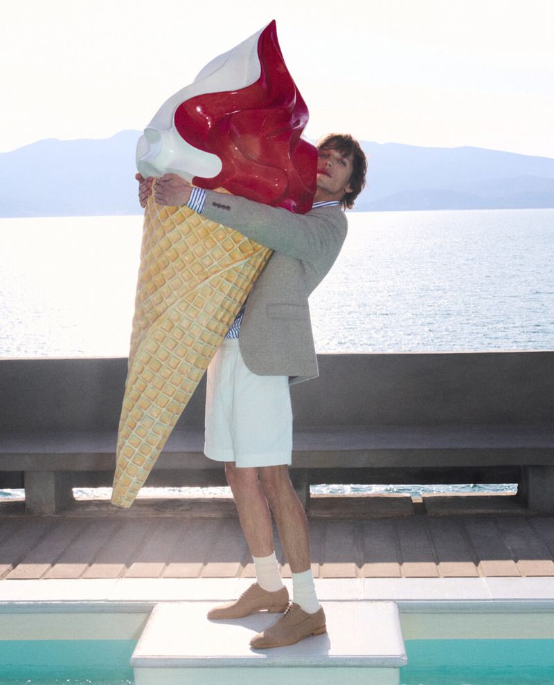 Man in a blazer and shorts stands on a white cube holding a giant inflatable ice cream cone with a lake and mountains behind him.