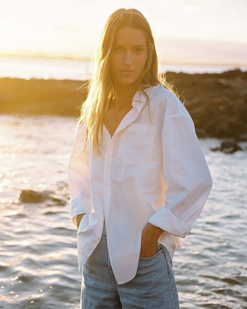 Woman in white button-down shirt stands near ocean at sunset, hands in pockets, blonde hair blowing in the wind, golden hour lighting.