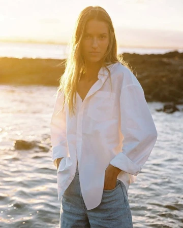 Woman in white button-down shirt stands near ocean at sunset, hands in pockets, blonde hair blowing in the wind, golden hour lighting.