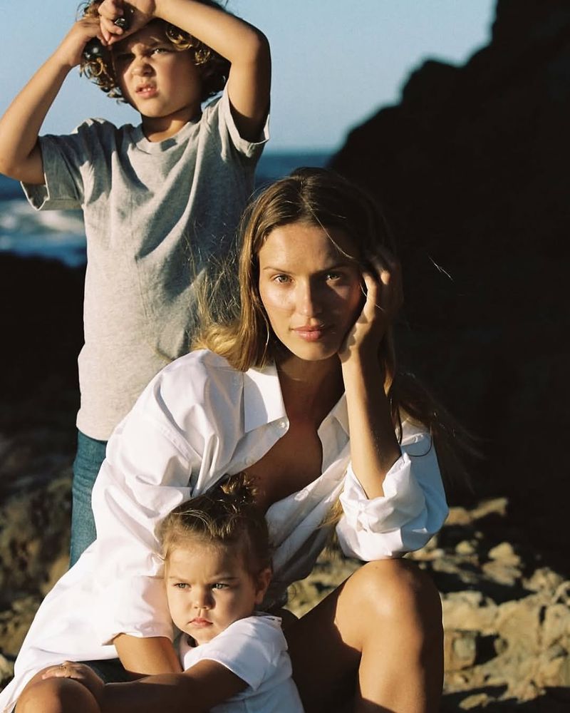 A sunlit outdoor portrait features a mother in a white shirt with her son and daughter, set against rocks and ocean, capturing a candid family moment.