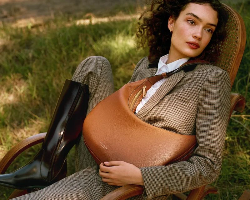 A woman in a tweed blazer and leather riding boot sits on a wood chair in a grassy field with a brown leather hobo bag.