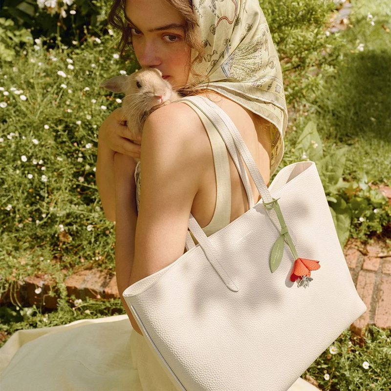 Woman sits in garden with a rabbit on her shoulder and a handbag, wearing a printed headscarf, dress, floral purse charm.