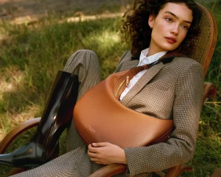 Model seated in a wooden chair wearing a tweed blazer and holding a brown leather shoulder bag outdoors