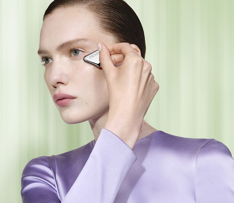 Close-up beauty shot of fair-skinned woman applying triangular, metallic Prada skincare tool to face in front of light green backdrop.