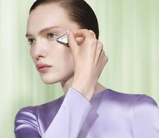 Close-up beauty shot of fair-skinned woman applying triangular, metallic Prada skincare tool to face in front of light green backdrop.