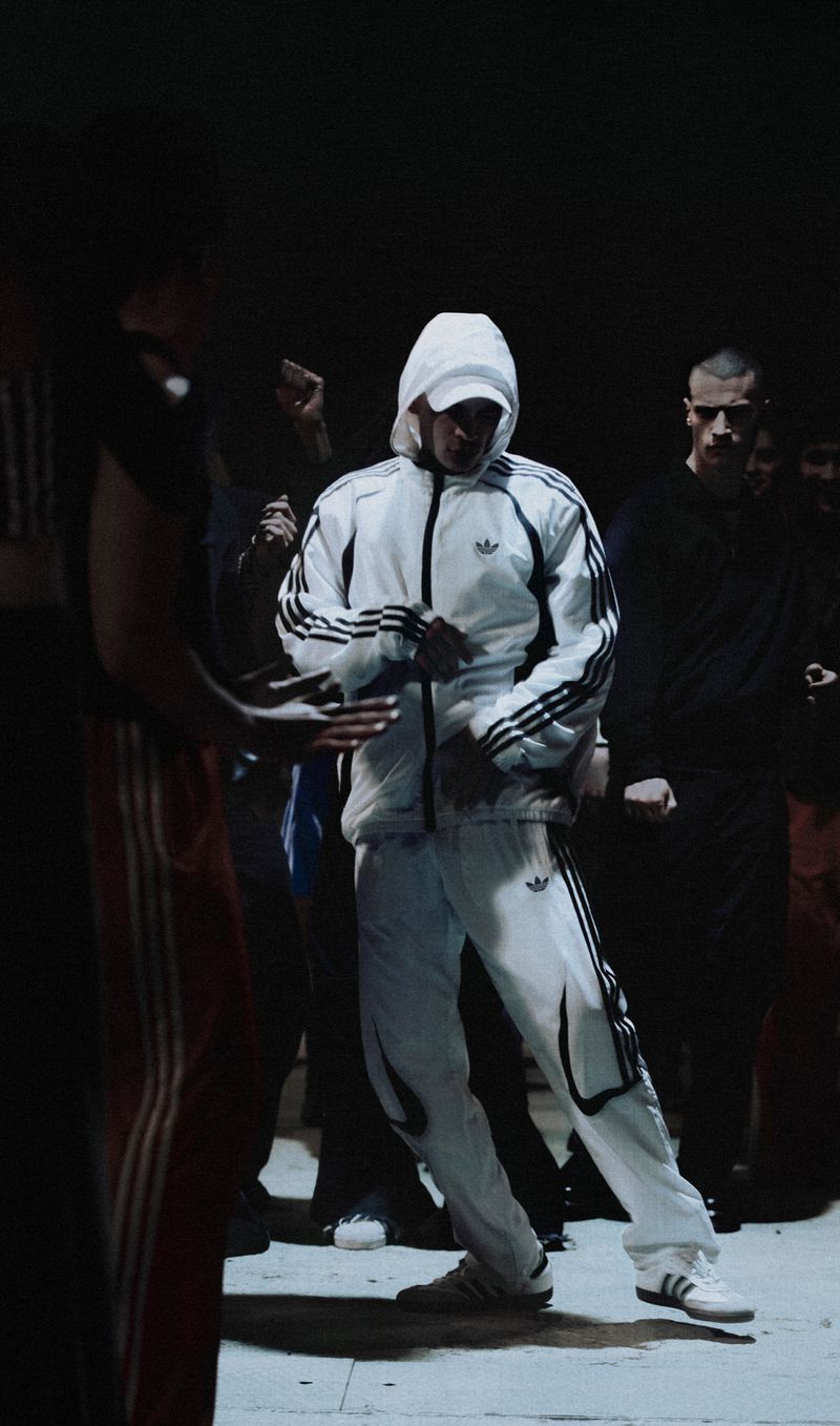 Man in a white tracksuit with black stripes dances in a dark urban setting, surrounded by other people, creating a candid street style scene.