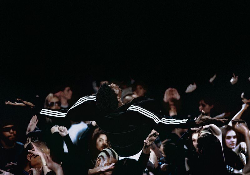 A crowd of people with hands raised, one man in a black Adidas track jacket is being held up above them, dark color palette.