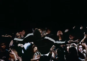 Man crowd surfing wearing a black Adidas track jacket with white stripes above raised hands in a dark venue