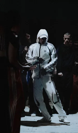 Man wearing a white Adidas tracksuit and hoodie dancing centrally in a group setting with low-key lighting