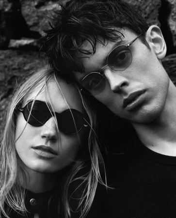 Black and white portrait of a stylish young couple wearing sunglasses posing in front of a textured dark stone wall outside.