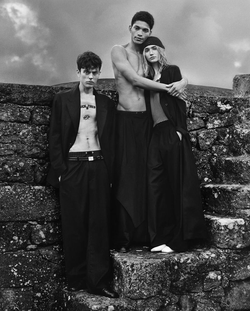 Three models in black clothing pose against a stone wall under a cloudy sky. The image is monochromatic with a fashion editorial style.