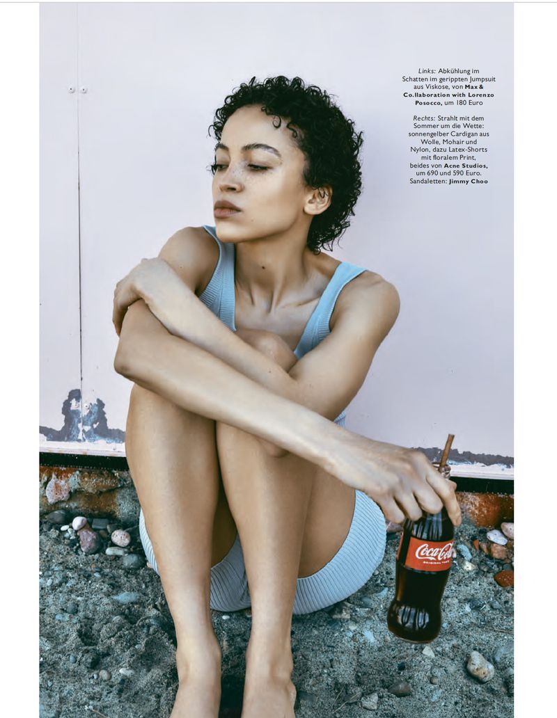 Young adult woman poses in fashion shot with a Coca-Cola bottle, sitting on stones against a weathered pink wall in a studio.