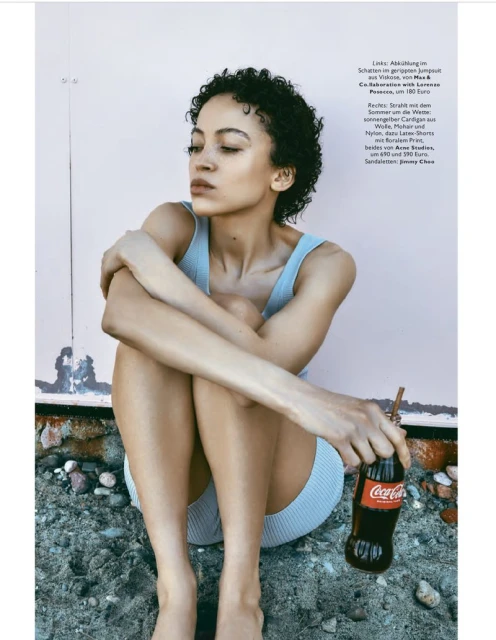 Young adult woman poses in fashion shot with a Coca-Cola bottle, sitting on stones against a weathered pink wall in a studio.