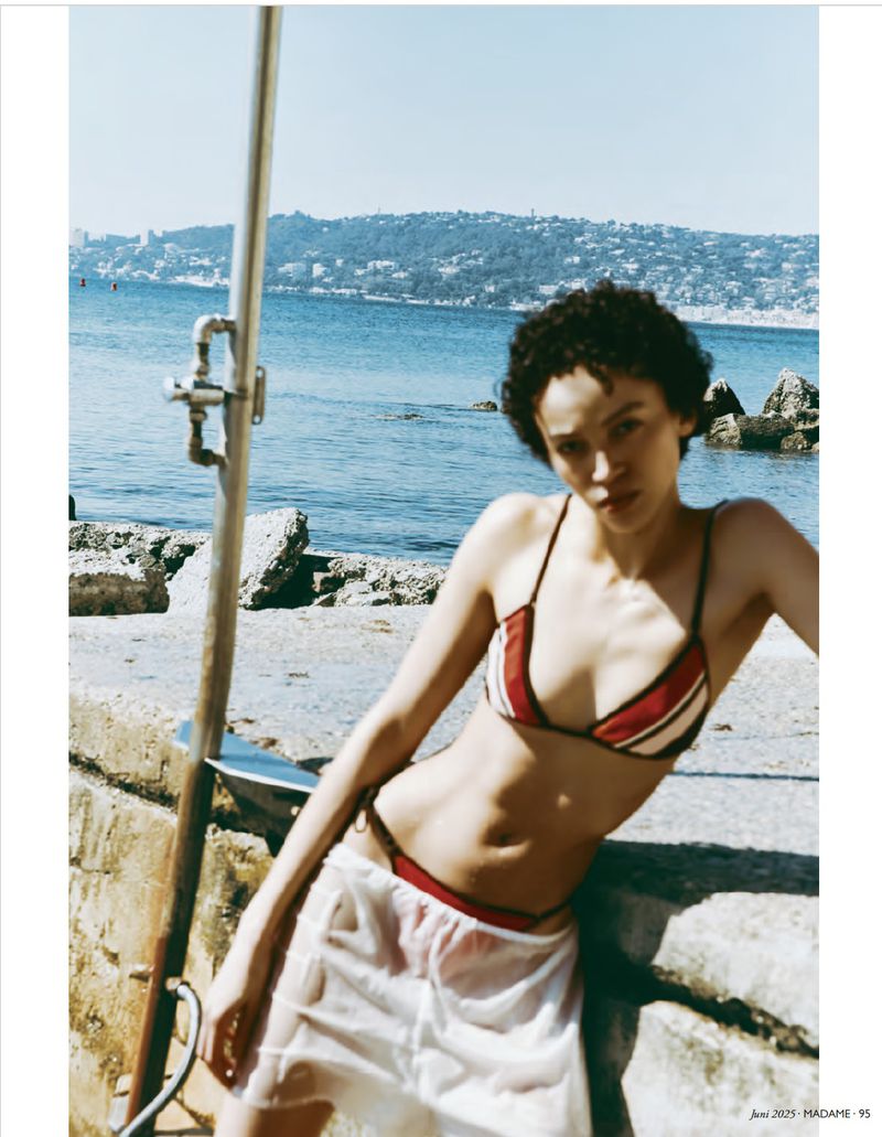 Woman poses in a bikini and cover-up by the ocean near a shower, capturing a fashion shot with coastal scenery and natural lighting.