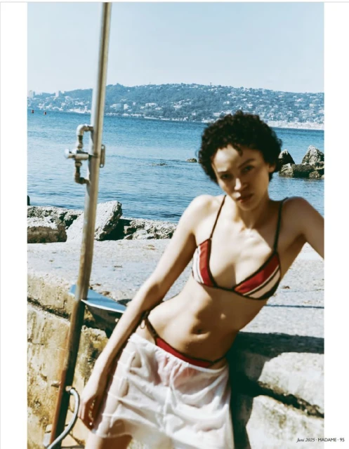 Woman poses in a bikini and cover-up by the ocean near a shower, capturing a fashion shot with coastal scenery and natural lighting.