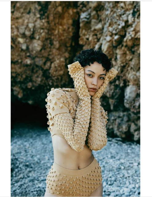 A tan-skinned model with curly dark hair poses on a stone beach. She is wearing a tan knit two-piece outfit with raised embellishments.