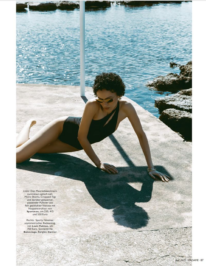 Woman in black swimsuit poses near ocean. Fashion editorial shot with concrete platform, rocks, and clear blue sea backdrop.