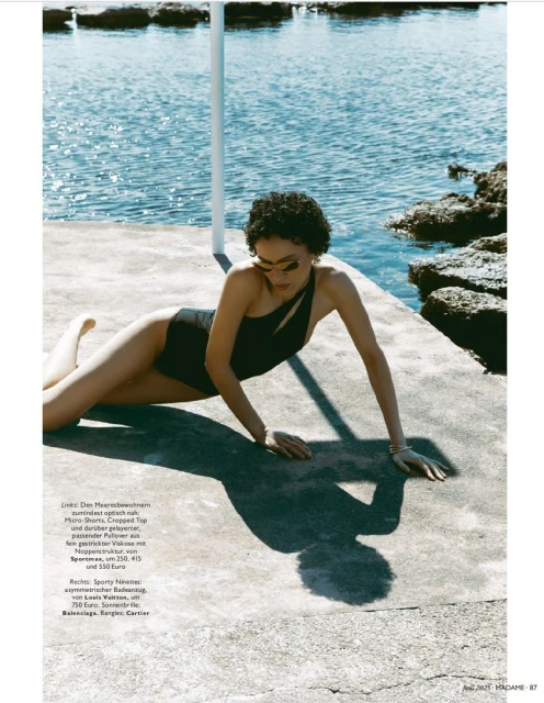Woman in black swimsuit poses near ocean. Fashion editorial shot with concrete platform, rocks, and clear blue sea backdrop.