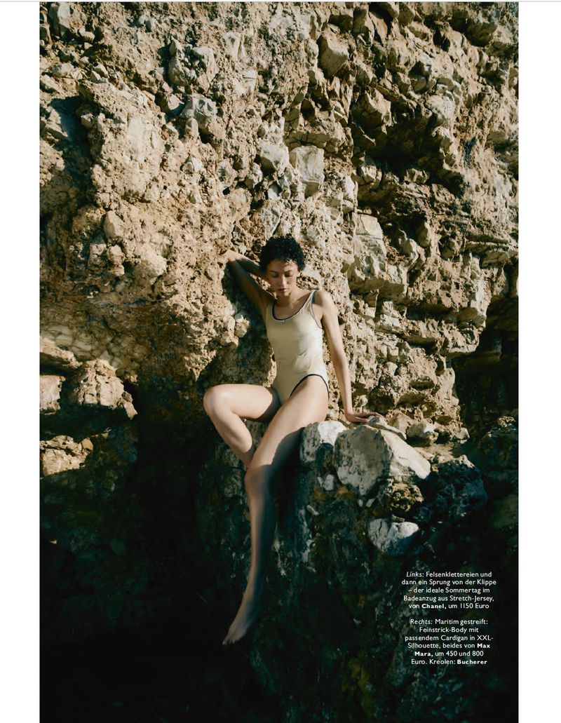 Woman in a cream-colored one-piece swimsuit poses on a rocky cliff near the coast, creating a fashion editorial vibe in natural light.