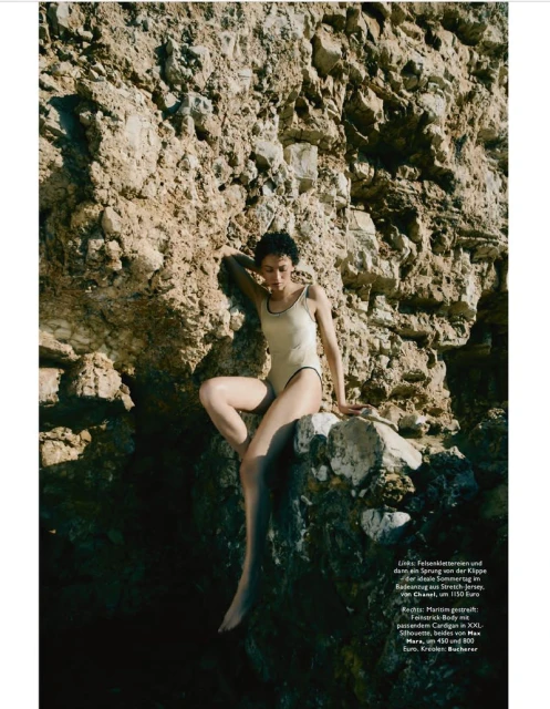 Woman in a cream-colored one-piece swimsuit poses on a rocky cliff near the coast, creating a fashion editorial vibe in natural light.