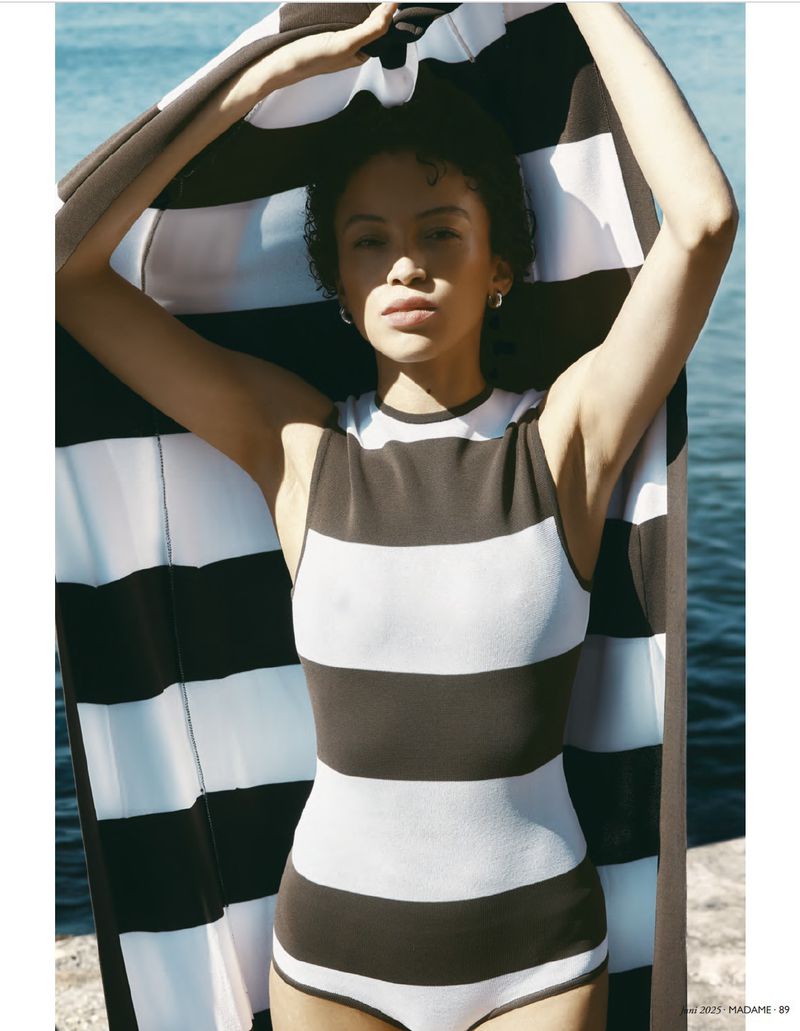A woman poses outdoors in a striped swimsuit holding a striped towel above her head near the ocean, showcasing fashion and style.