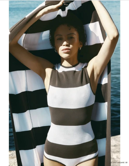 A woman poses outdoors in a striped swimsuit holding a striped towel above her head near the ocean, showcasing fashion and style.
