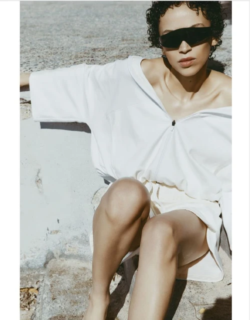 Woman with short curly hair wears sunglasses and an oversized white shirt while sitting on a stone surface in an outdoor setting.