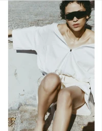 Model wearing dark visor sunglasses and white outfit seated outdoors against textured stone wall