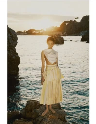 Model standing on coastal rocks at sunset wearing a white top and yellow pleated skirt overlooking the sea