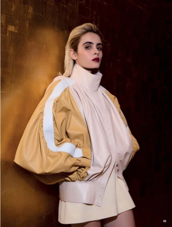 Fashion portrait of a blonde adult woman in a studio setting wearing a tan jacket, looking at the camera with a neutral expression.