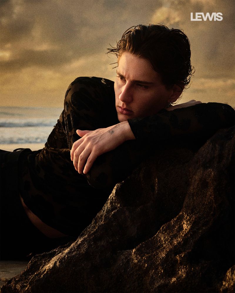 Portrait of a male model with dark hair reclining on rocks near a beach, wearing a black top, against a sunset-toned sky with waves.