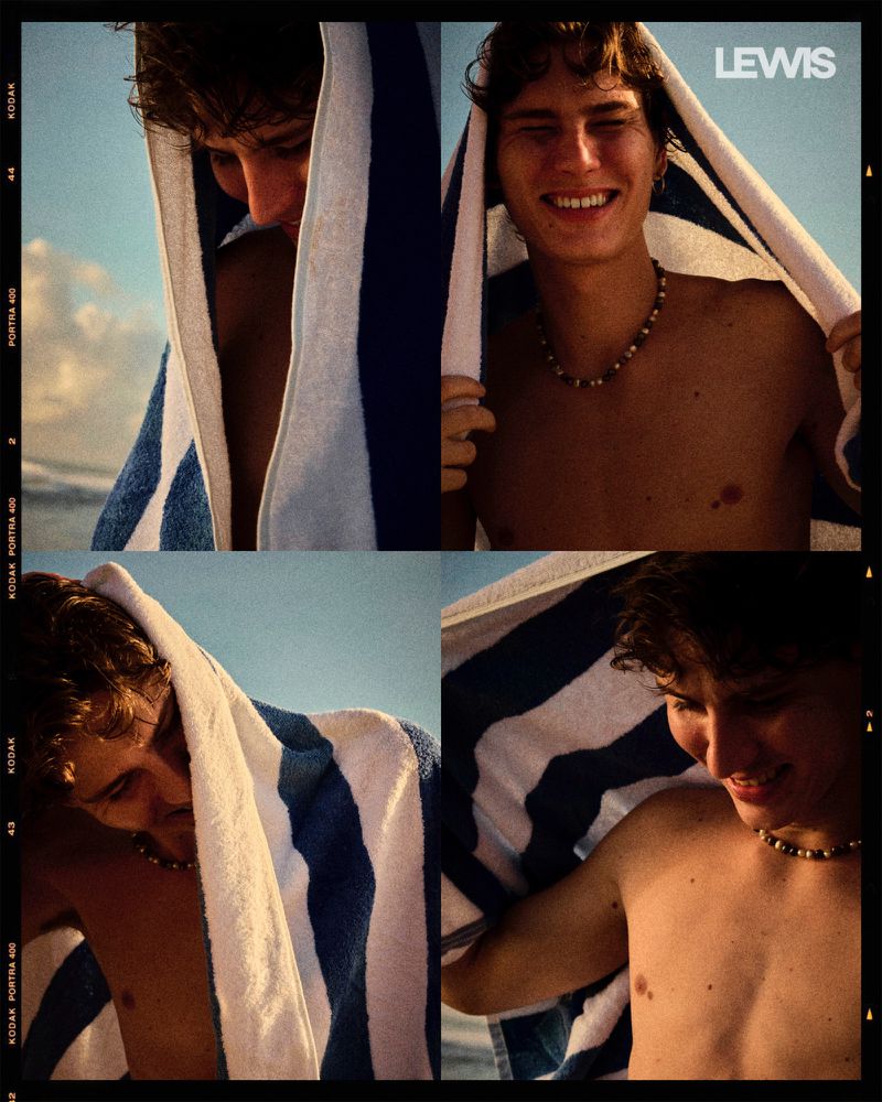 Portrait of a smiling young adult male with a beaded necklace, wrapped in a blue and white striped towel, captured in a bright outdoor setting.