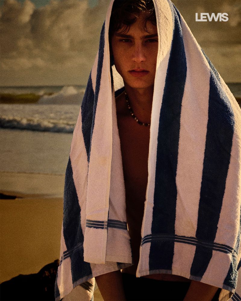 A young adult male wrapped in a blue-and-white striped towel stands on a beach, with ocean waves visible in the background.