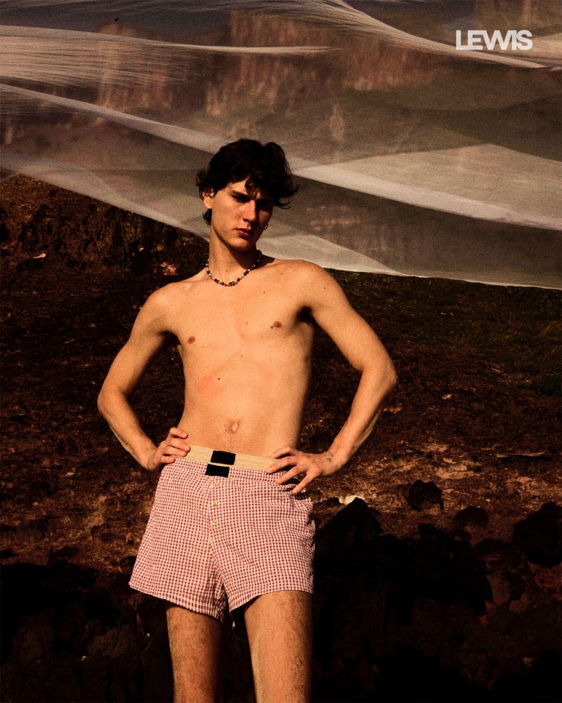 Young adult man poses shirtless outdoors in red and white checkered shorts with a textured background, in soft, warm lighting.