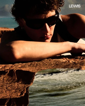 A young adult male with sunglasses rests on a rock formation overlooking ocean waves in this lifestyle-oriented outdoor portrait.