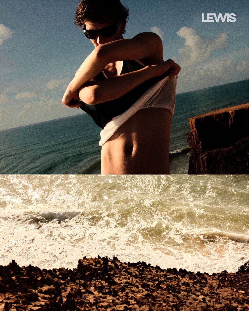 Man on cliffside removes shirt overlooking sea. Features blue sky, waves crashing on rocks, with 'Lewis' logo in corner, in warm light.