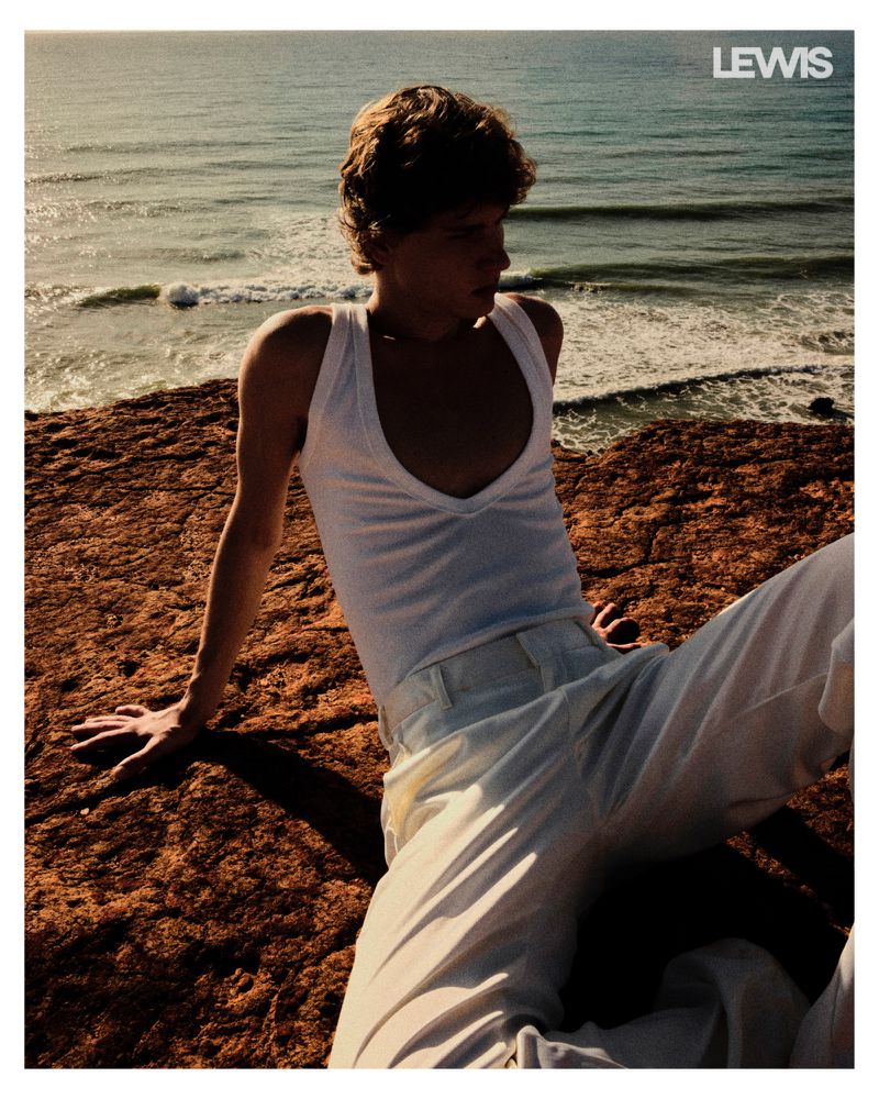 Young adult male poses on rocky shore with ocean backdrop, wearing a white tank top and pants, lit by warm sunlight in outdoor setting.